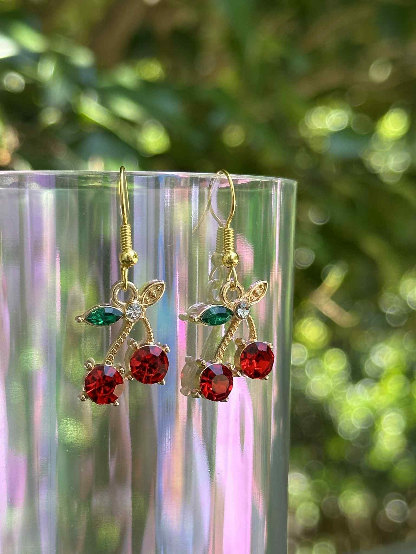 Gold earrings with red and green gemstones on a clear stand with a blurred green background