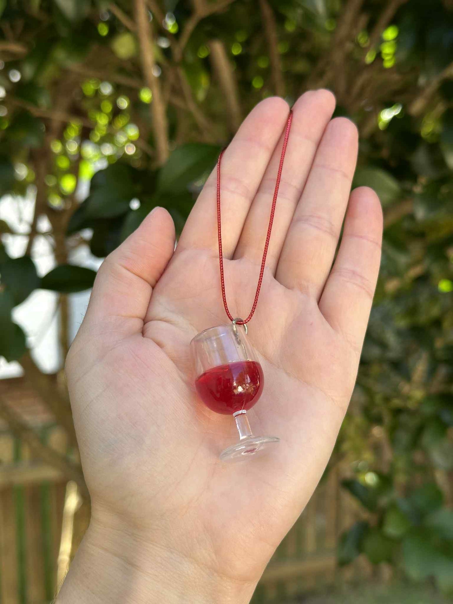 Hand holding a small glass charm with red liquid against a blurred green outdoor background