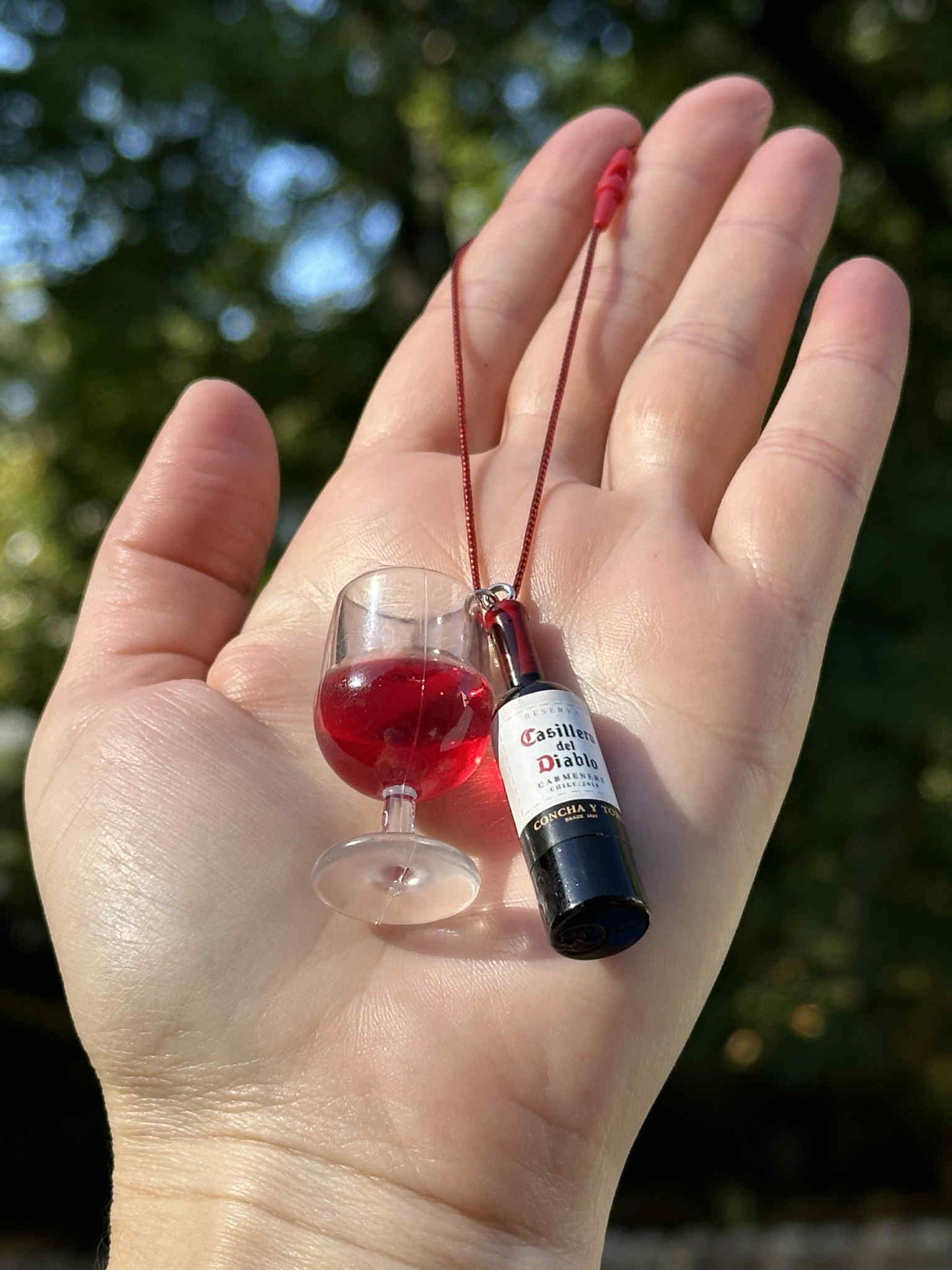 Miniature red drink bottle and glass ornament with hanging cord, held in hand outdoors.