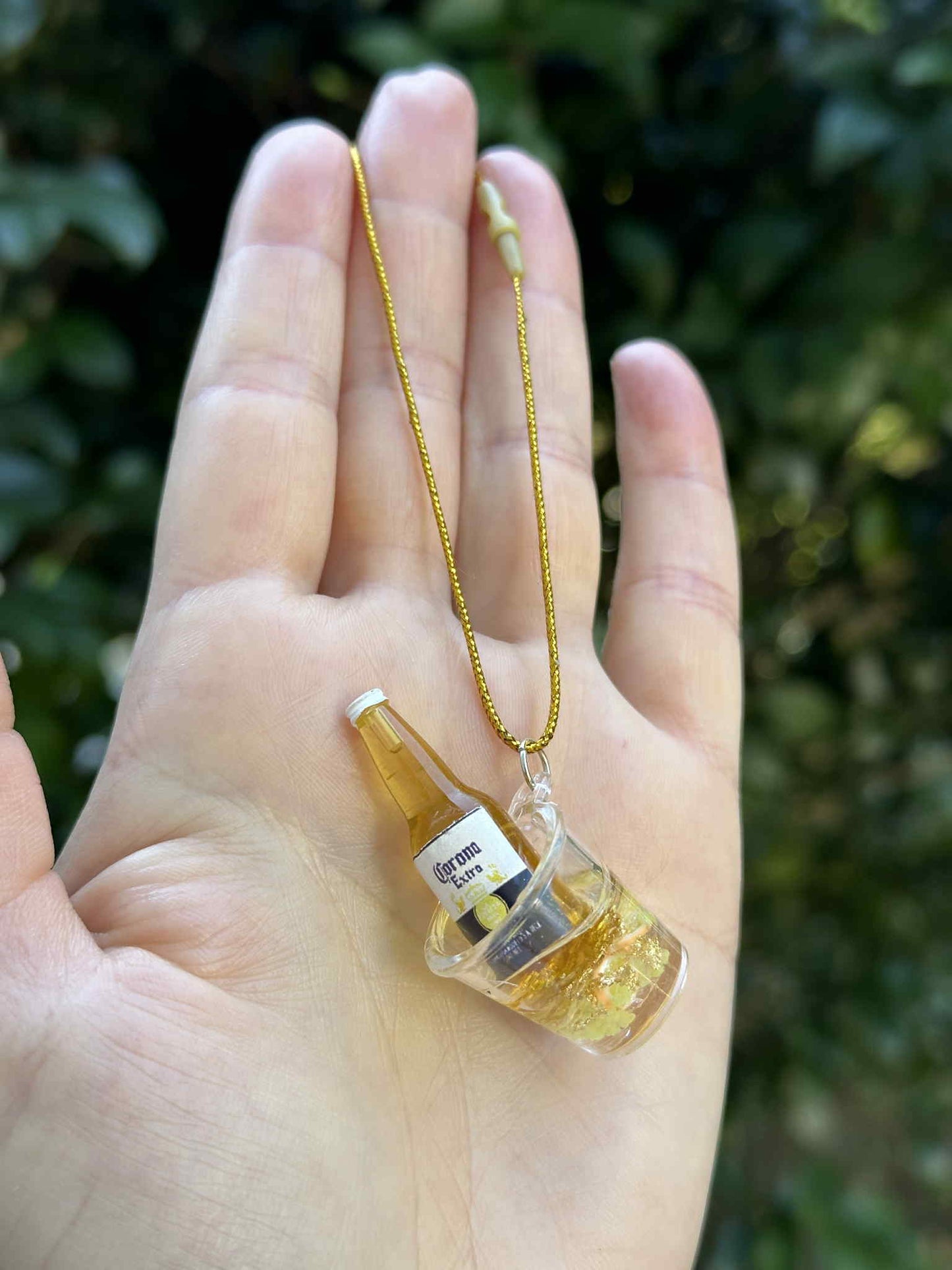 Miniature golden bottle in ice bucket ornament with gold hanging cord, held in hand outdoors.