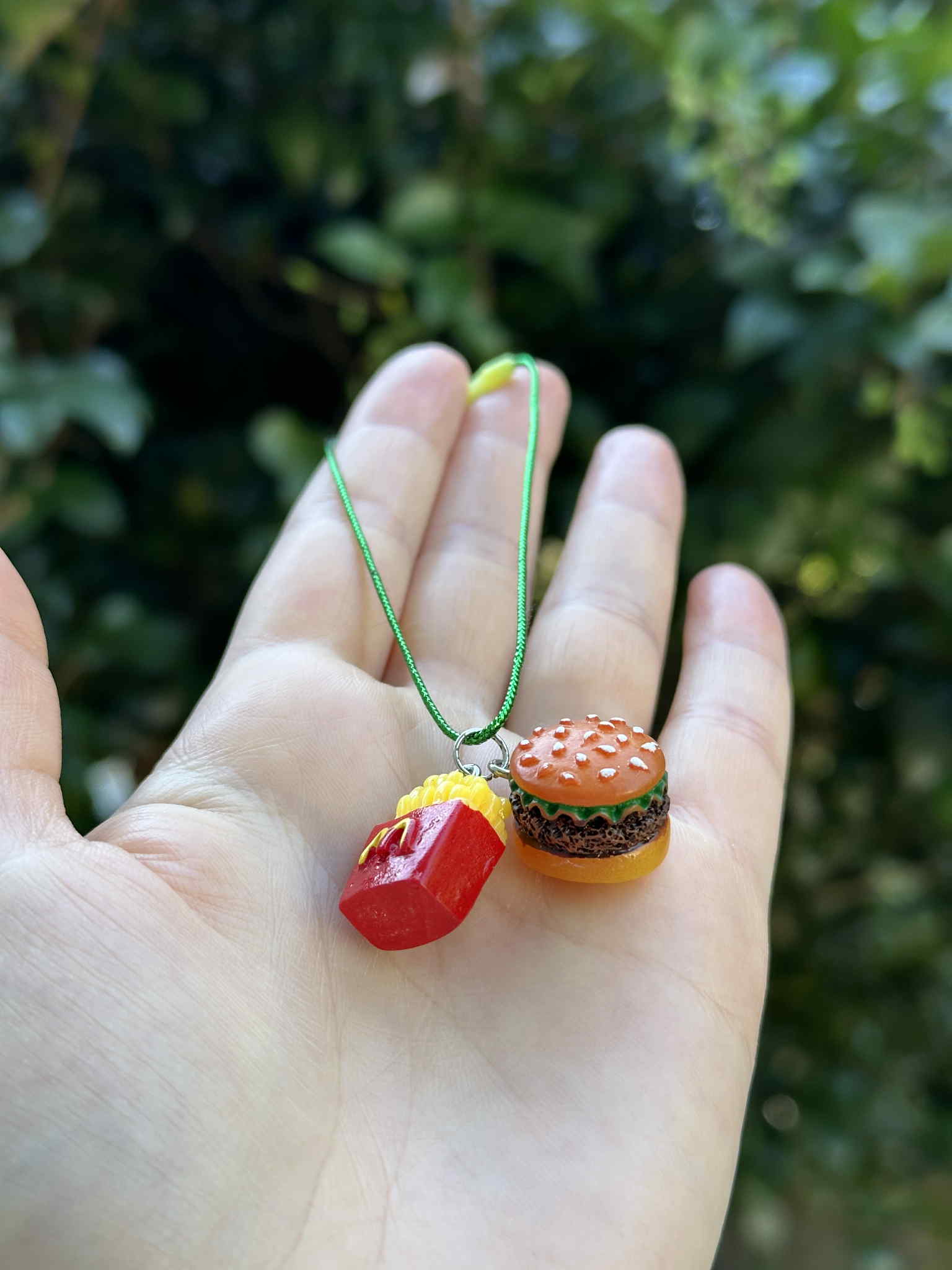 Mini burger and fries ornament with green hanging cord, held in hand outdoors.