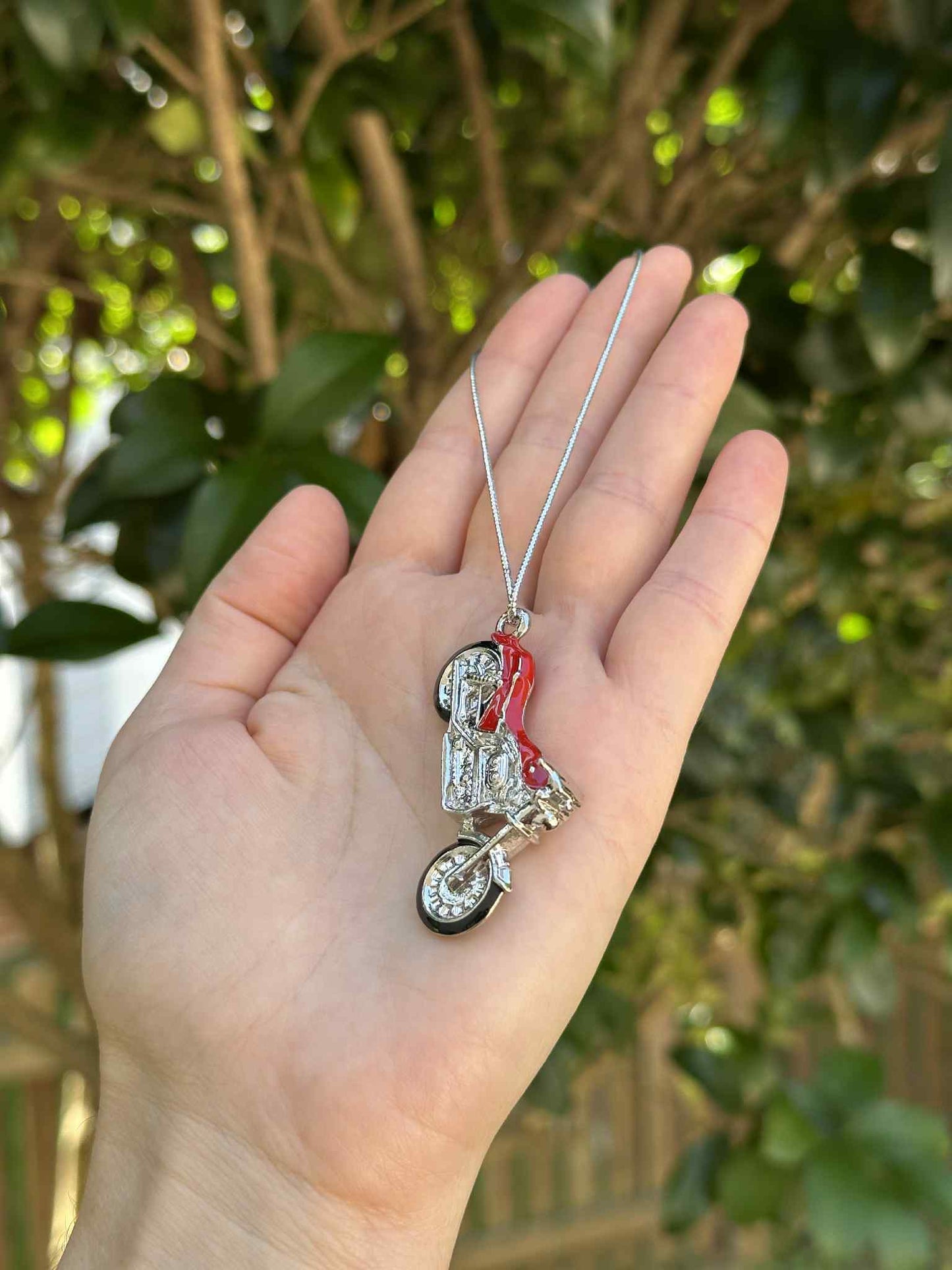 Hand holding a necklace with a baby carriage pendant against a blurred green leafy background
