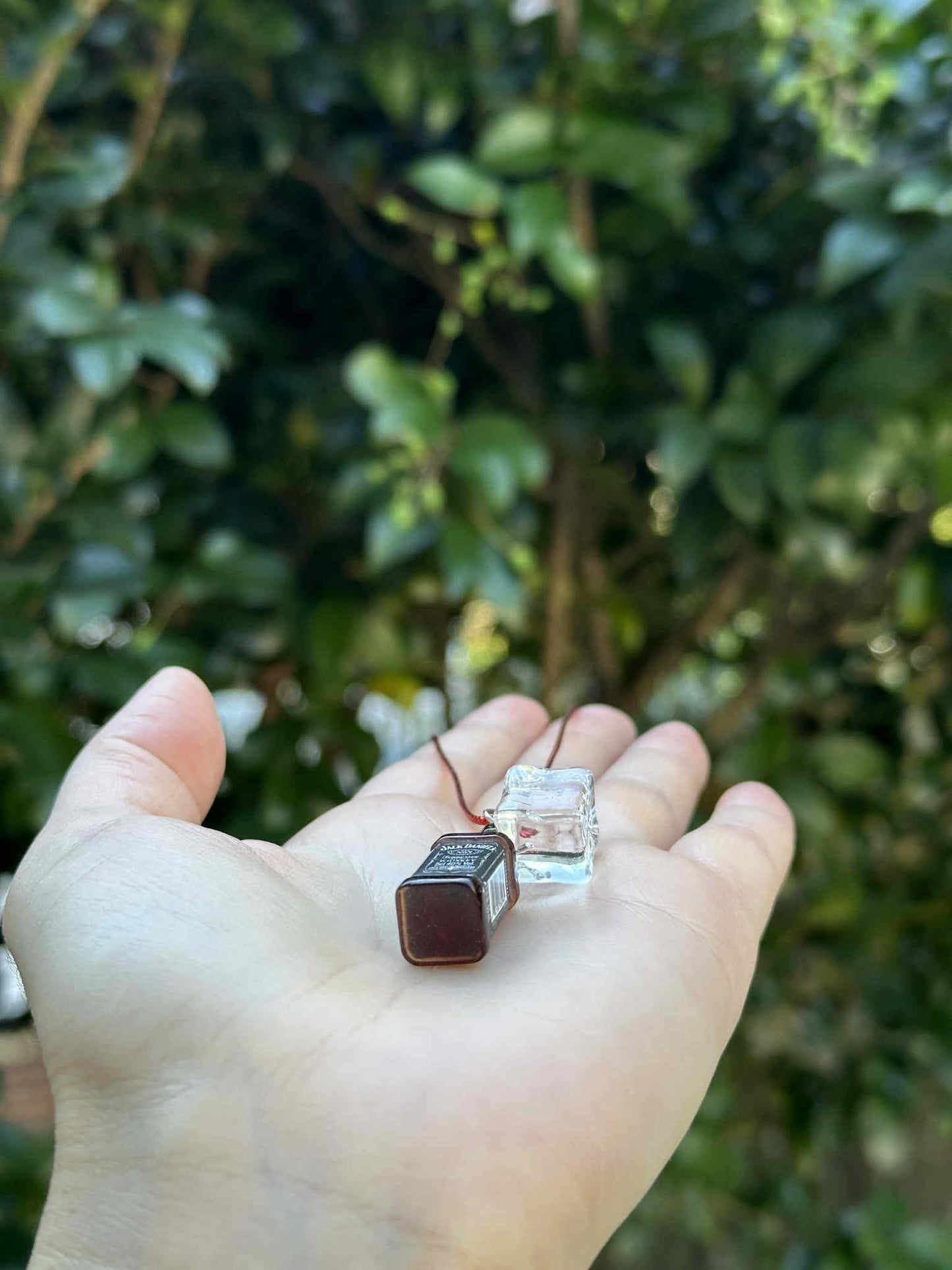 Miniature brown bottle and clear glass ornament with red hanging cord, held in hand outdoors.