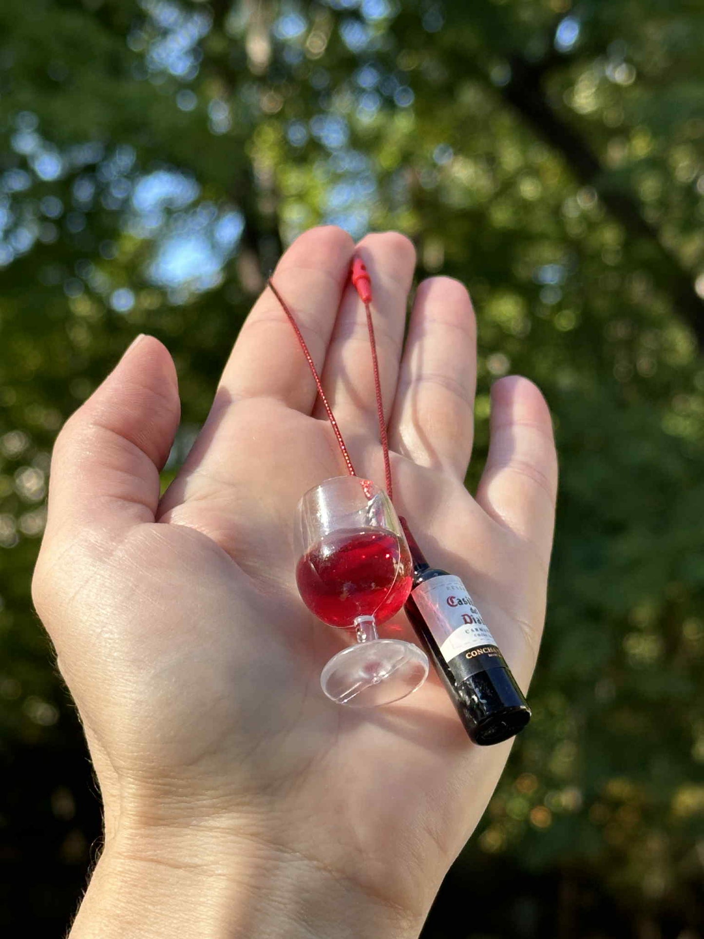 Miniature red drink bottle and glass ornament with hanging cord, held in hand outdoors.