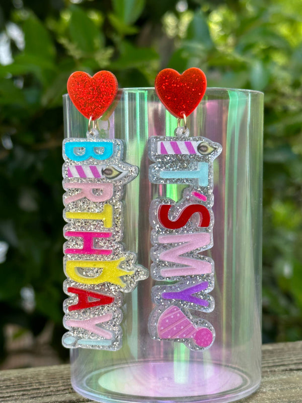Colorful earrings with heart charms on a clear stand against a blurred green background