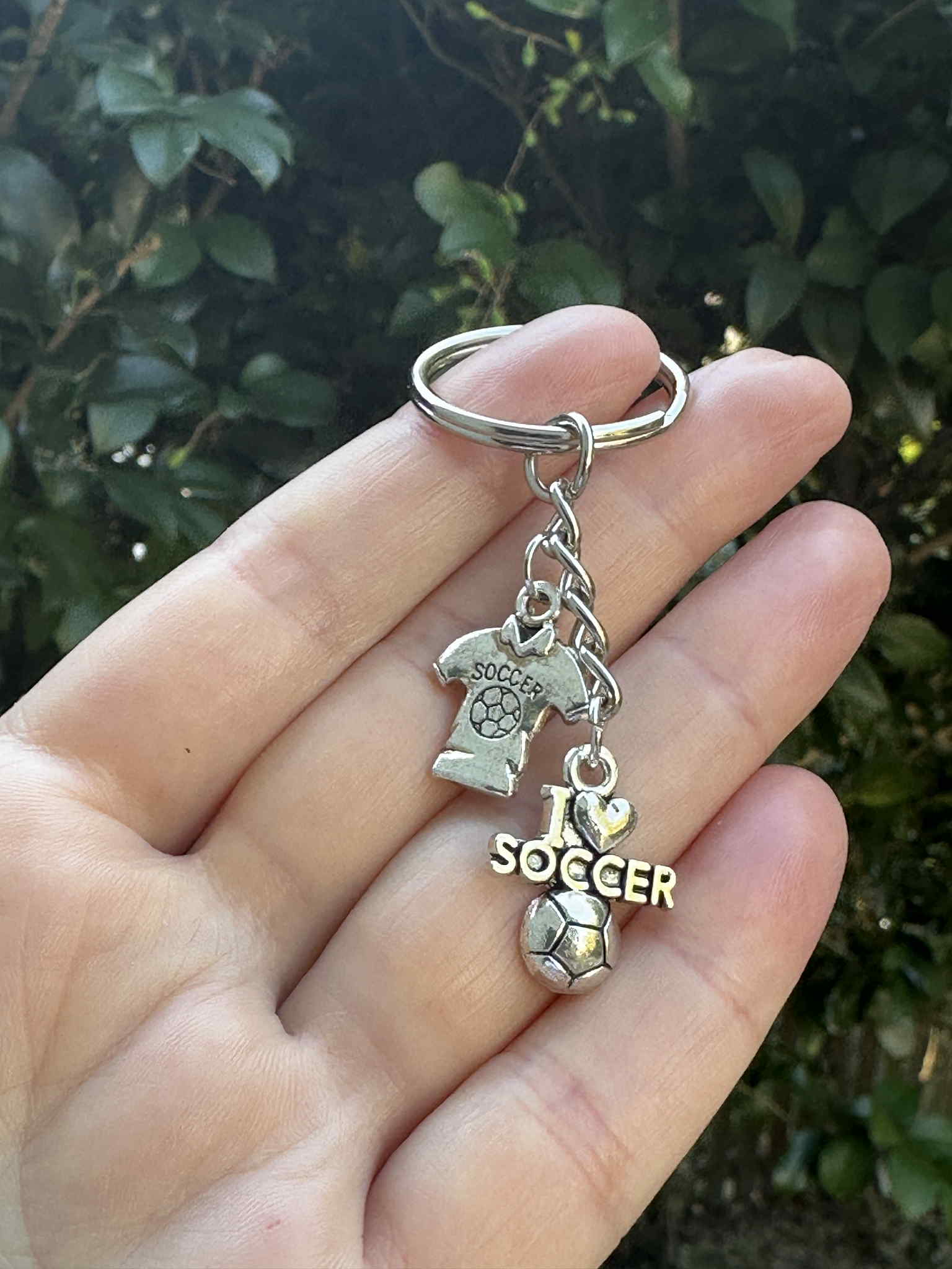 Silver soccer keychain with I Love Soccer charm, jersey pendant, and soccer ball charm held in hand outdoors.