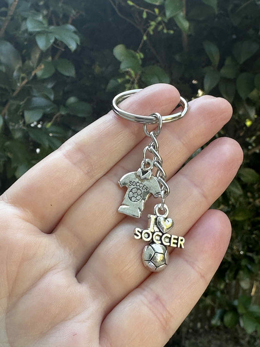Silver soccer keychain with I Love Soccer charm, jersey pendant, and soccer ball charm held in hand outdoors.