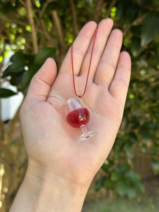 Hand holding a small glass pendant with red liquid against a blurred green outdoor background