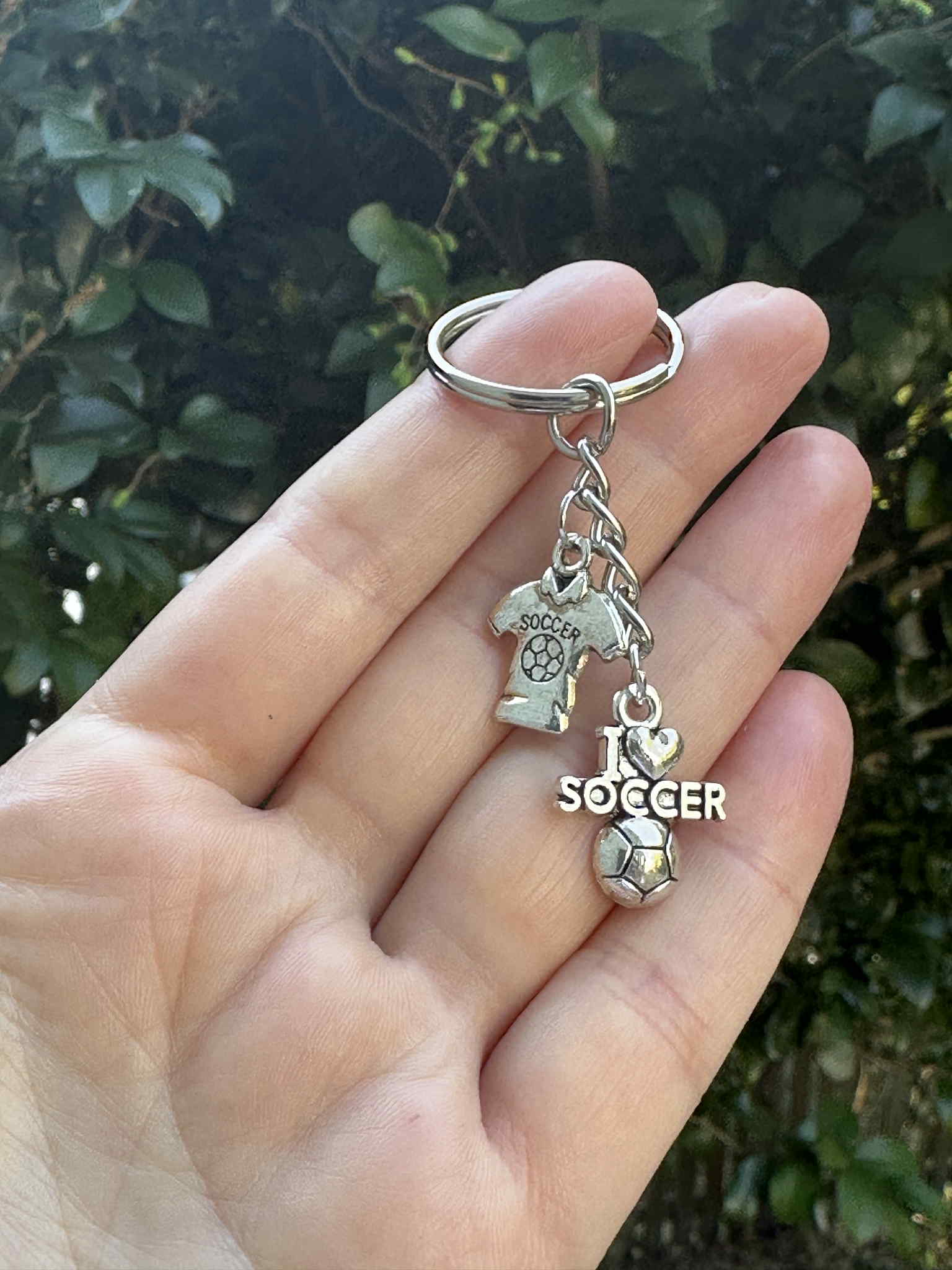 Silver soccer keychain with I Love Soccer charm, jersey pendant, and soccer ball charm held in hand outdoors.