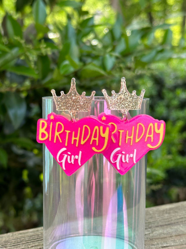 Clear cup with 'Birthday Girl' decorations featuring pink hearts and gold crowns against a blurred green outdoor background.