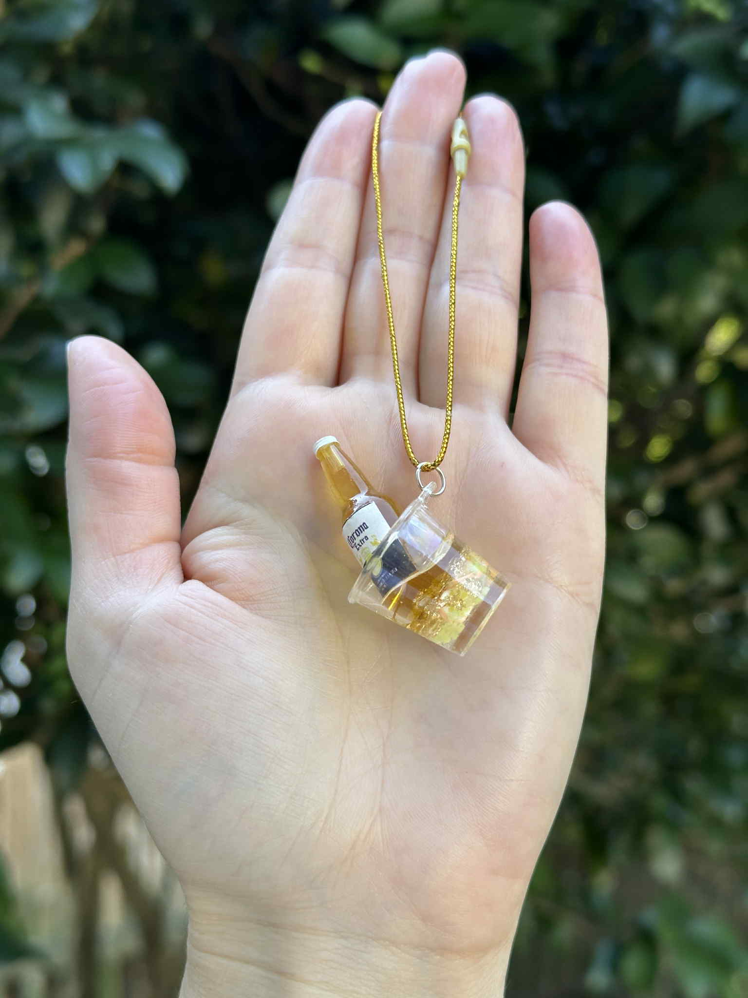 Miniature golden bottle in ice bucket ornament with gold hanging cord, held in hand outdoors.