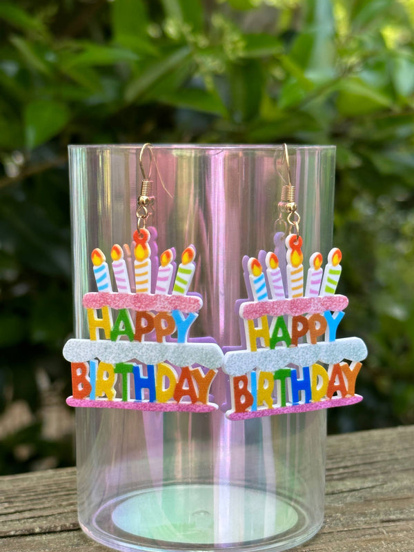 Colorful 'Happy Birthday' cake-shaped earrings in a clear display case with a green leafy background.