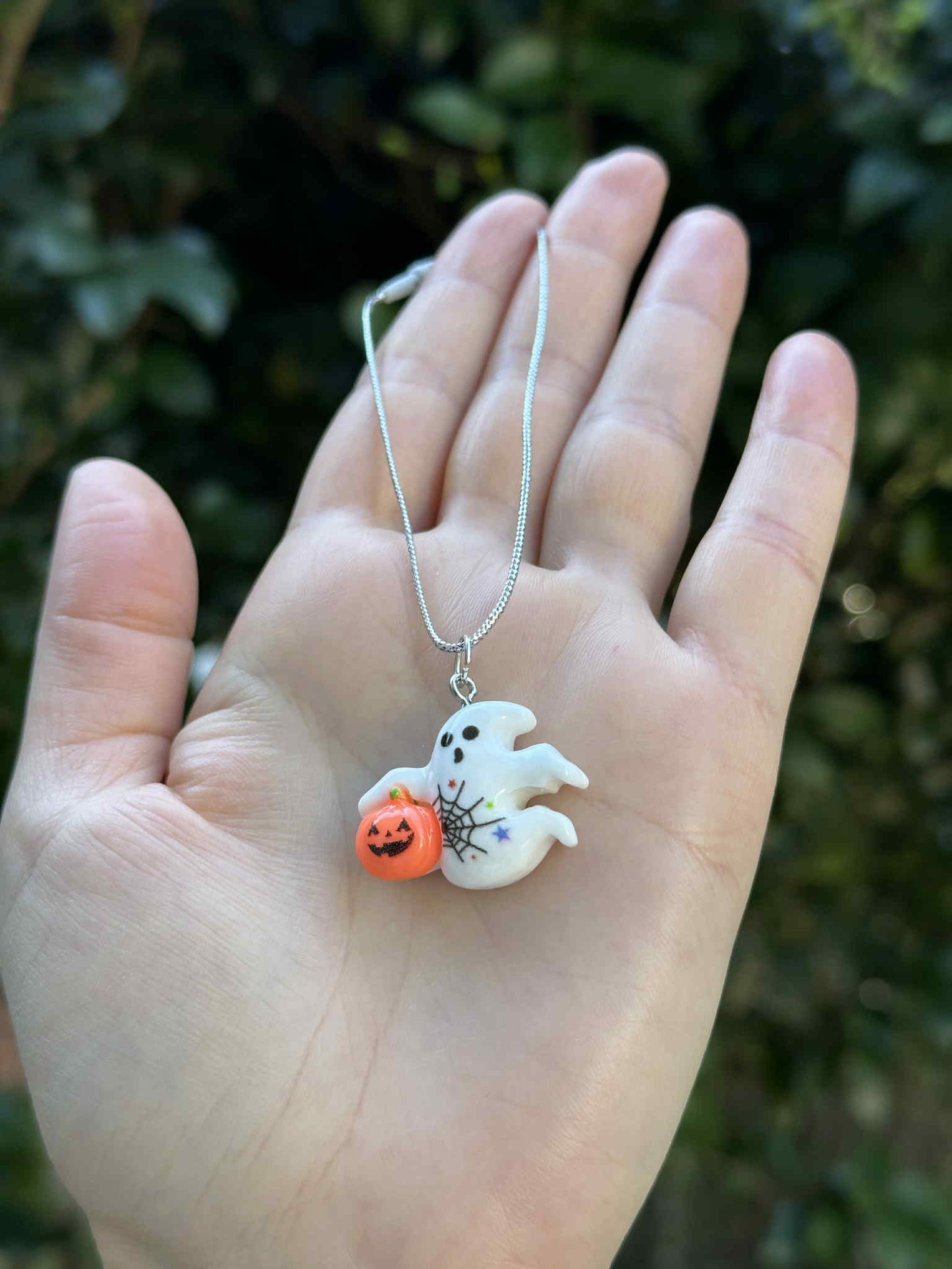 Mini ghost holding pumpkin ornament with silver hanging cord, Halloween decoration held in hand outdoors.