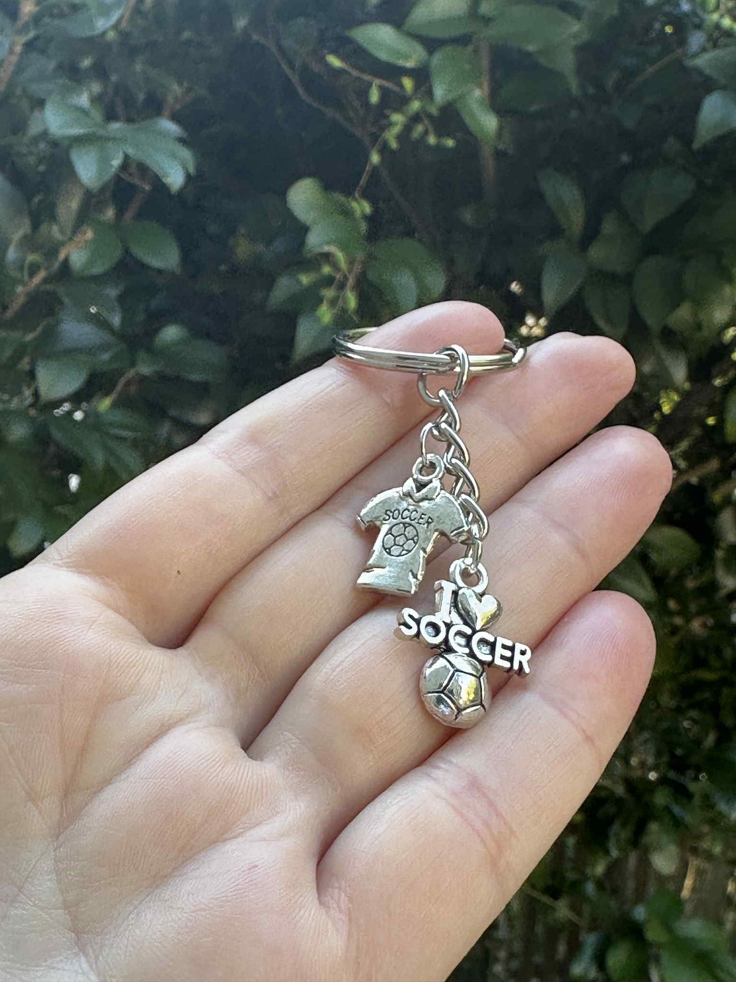 Silver soccer keychain with I Love Soccer charm, jersey pendant, and soccer ball charm held in hand outdoors.