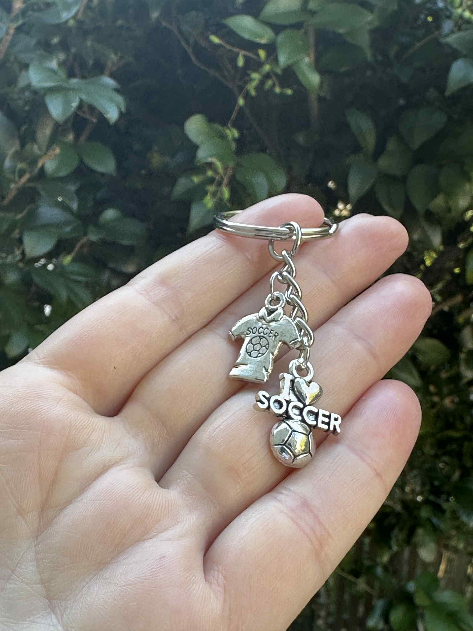 Silver soccer keychain with I Love Soccer charm, jersey pendant, and soccer ball charm held in hand outdoors.