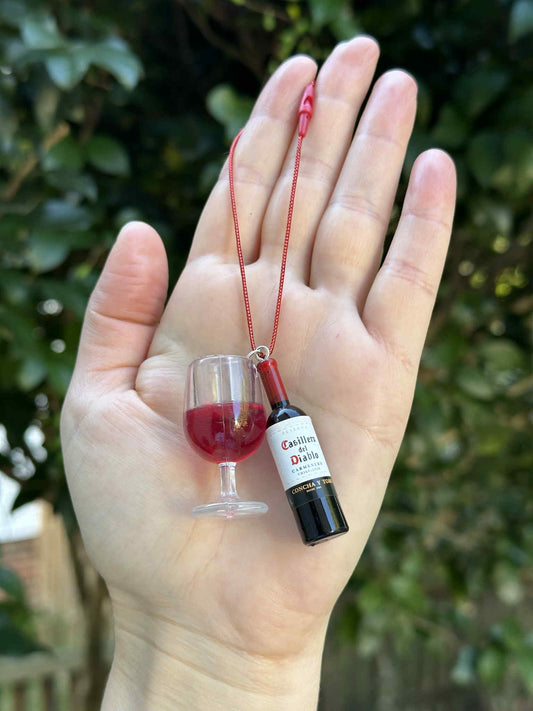 Miniature red drink bottle and glass ornament with hanging cord, held in hand outdoors.