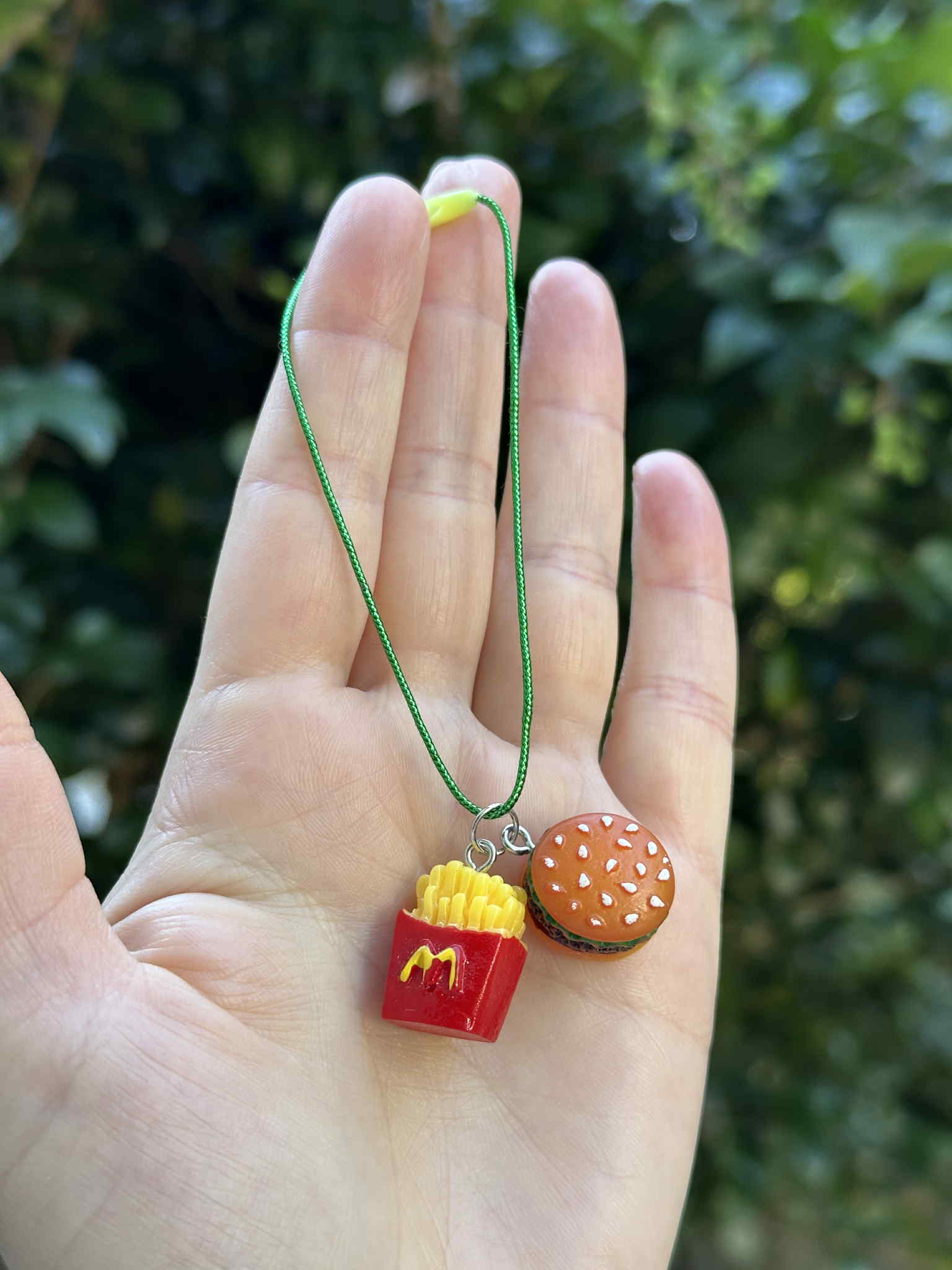 Mini burger and fries ornament with green hanging cord, held in hand outdoors.