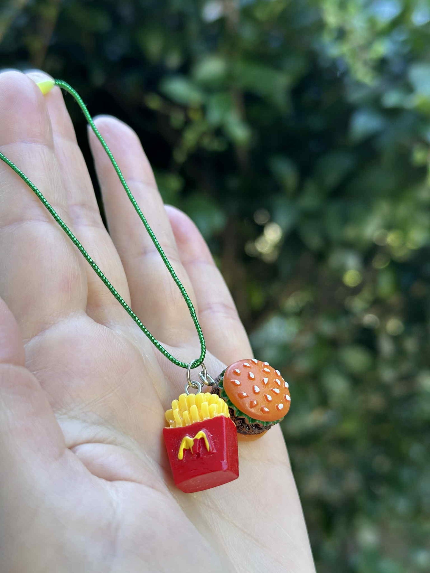Mini burger and fries ornament with green hanging cord, held in hand outdoors.