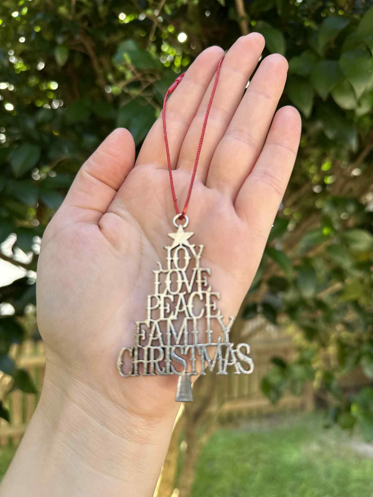 Hand holding a Christmas ornament with 'Joy Love Peace Family Christmas' text against a blurred green outdoor background.