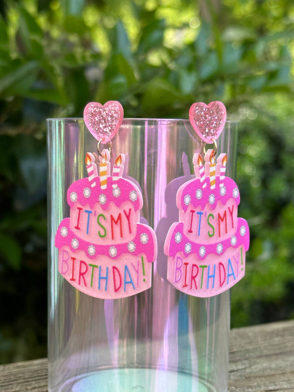 Pink 'It's My Birthday!' earrings with heart-shaped clips on a clear stand against a blurred green background.
