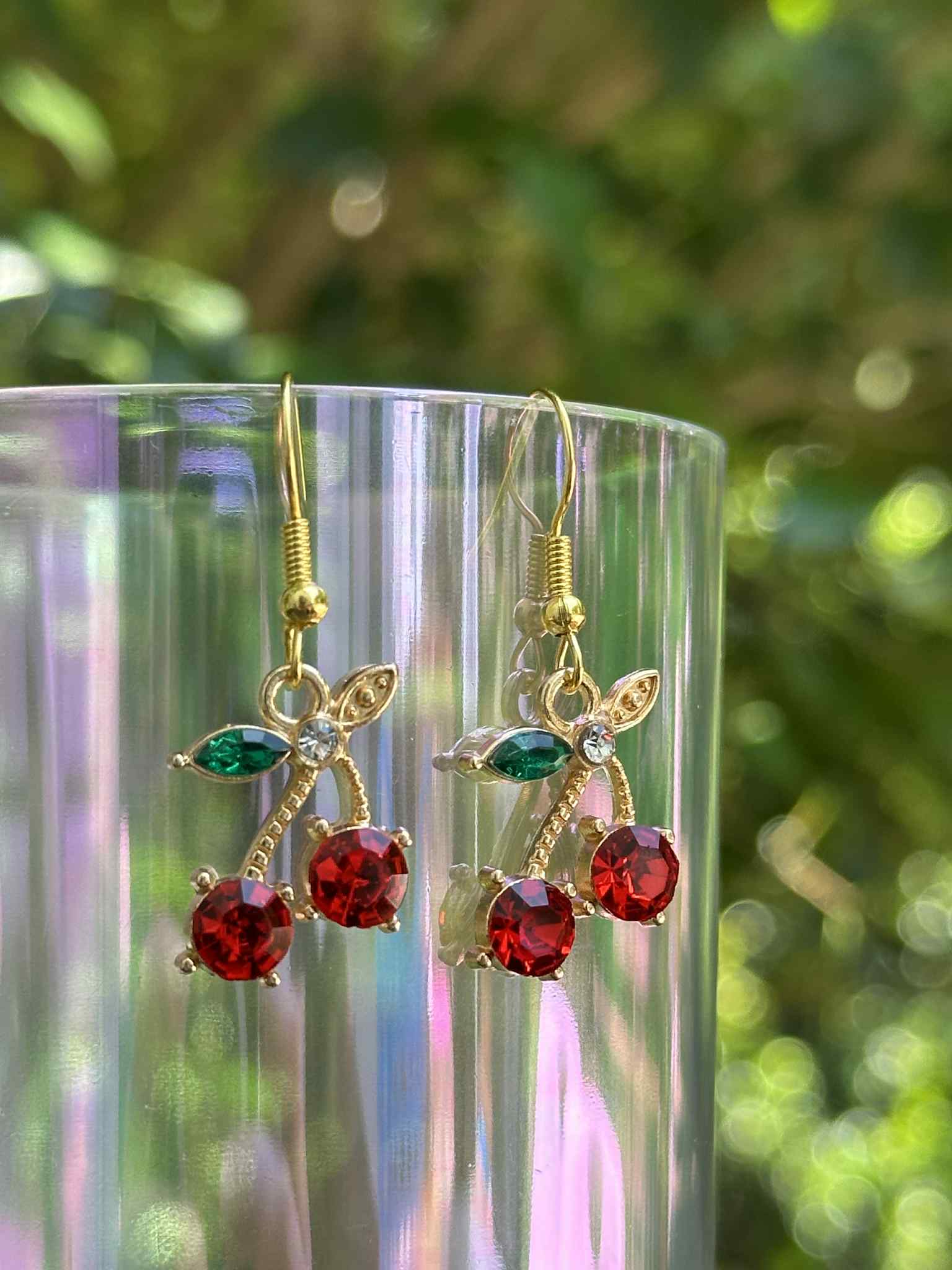 Gold earrings with red and green gemstones on a clear stand with a blurred green background