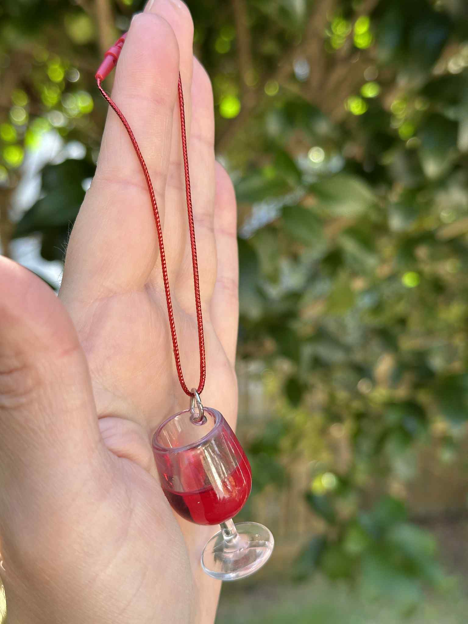 Hand holding a necklace with a glass pendant filled with red liquid against a blurred green background