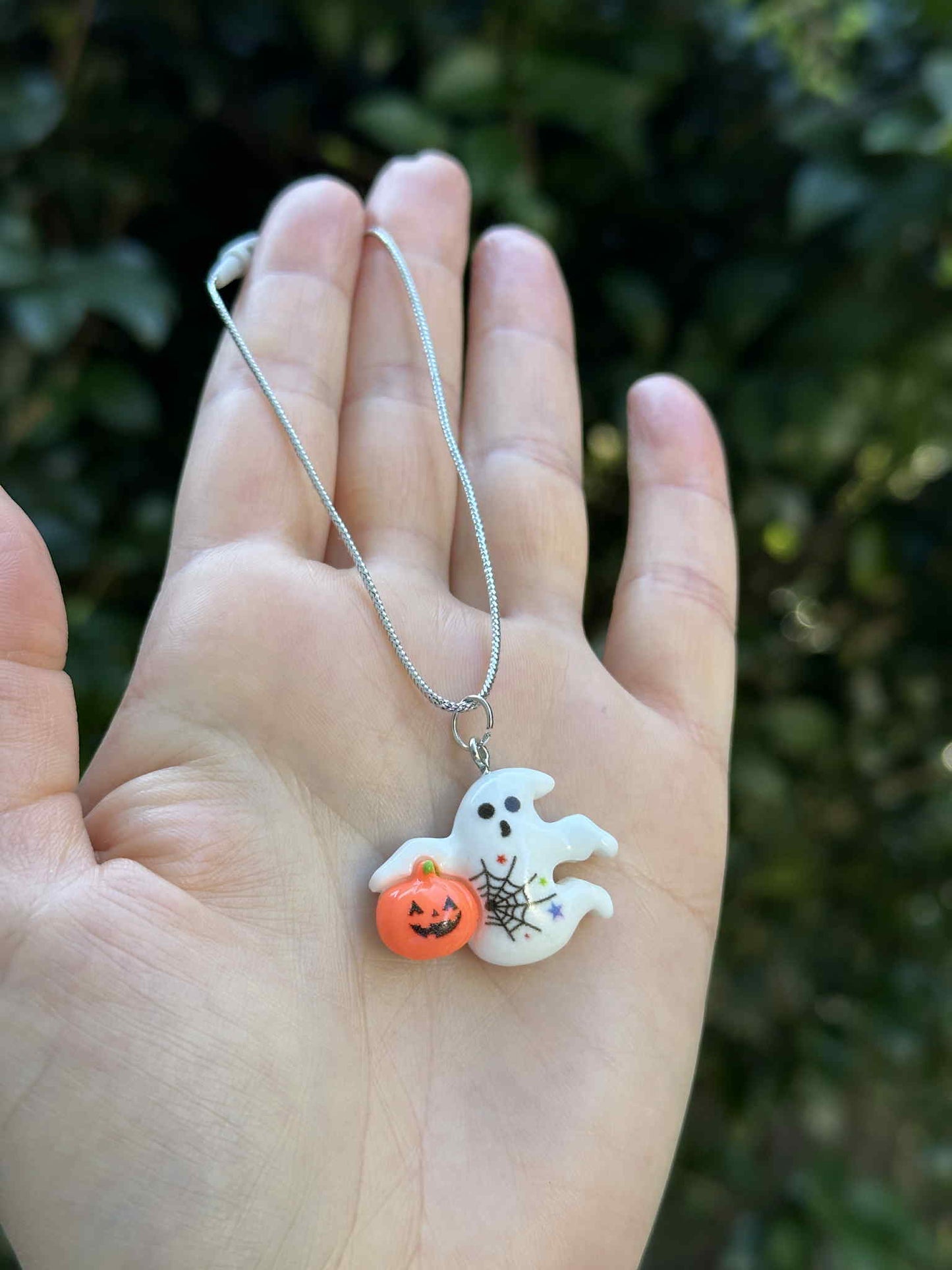 Mini ghost holding pumpkin ornament with silver hanging cord, Halloween decoration held in hand outdoors.