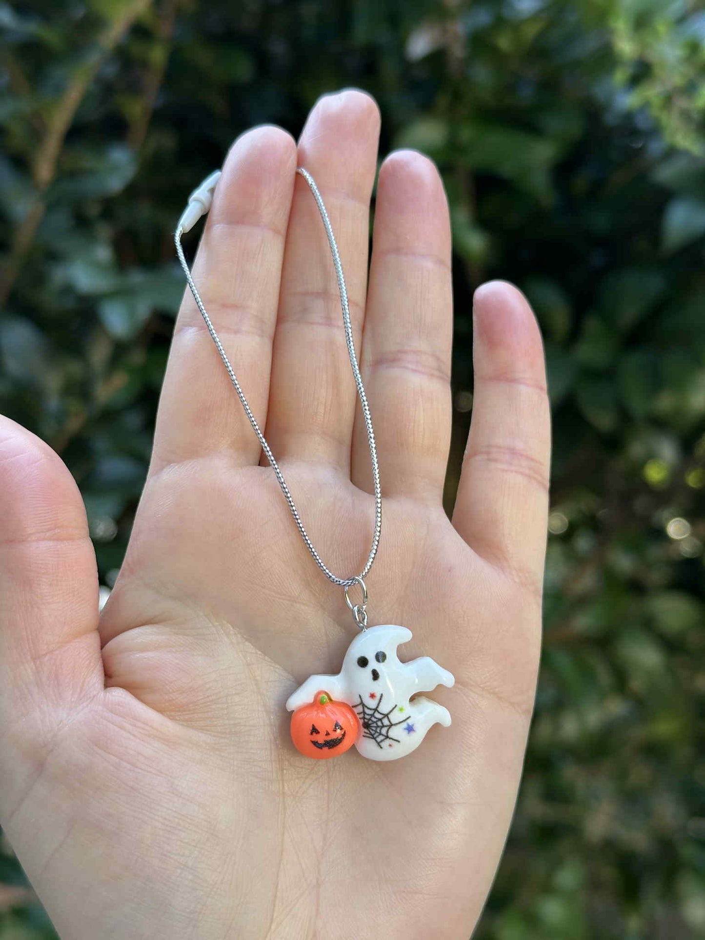 Mini ghost holding pumpkin ornament with silver hanging cord, Halloween decoration held in hand outdoors.