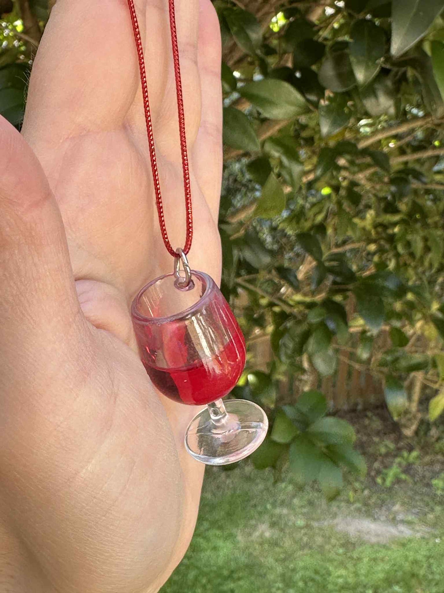 Hand holding a necklace with a glass pendant of a wine glass filled with red liquid, against a blurred green outdoor background.
